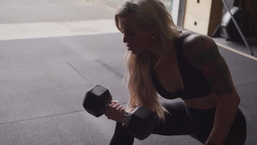 A woman with blonde hair and a tattooed arm is intensely focused while doing a bicep curl with a dumbbell