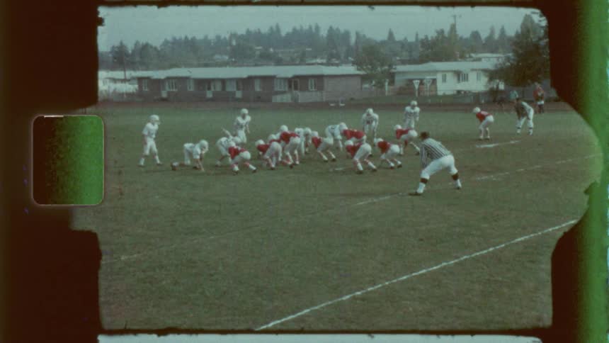 A group of young football players competes on a grassy field with one team in red jerseys the other in white and a referee observing nearby