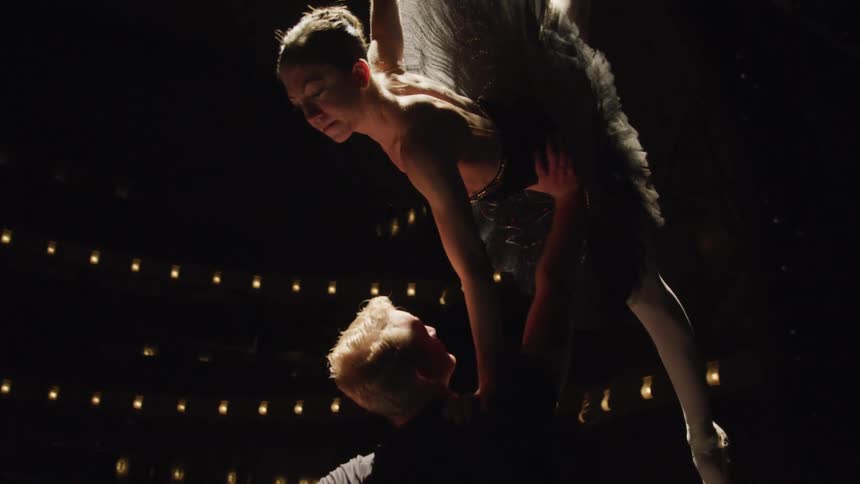 Male ballet dancer holds a ballerina above his head in a dark theater