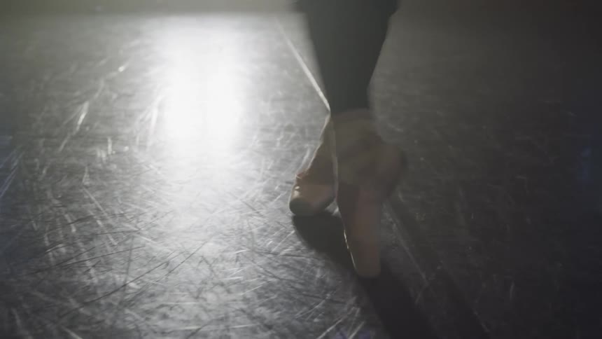 A closeup shows a dancers feet in worn ballet shoes on a scratched reflective dance floor illuminated by a bright light source