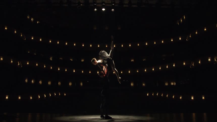 Two dancers on a lit stage with one of them being held by the other in an acrobatic pose