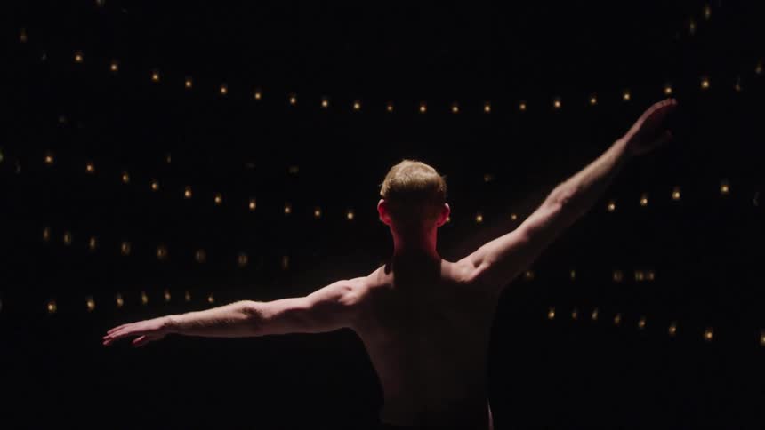 A shirtless ballet dancer with arms outstretched stands on a dark stage illuminated by rows of small lights in the background
