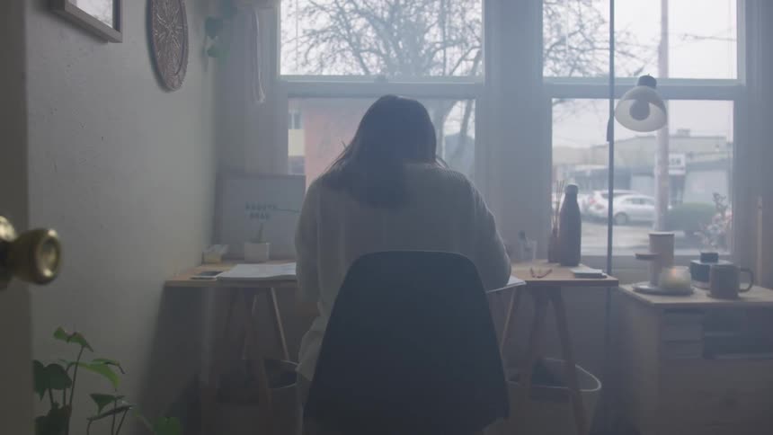 A person with long dark hair sits at a desk in a room with a large window overlooking a gray outdoor scene