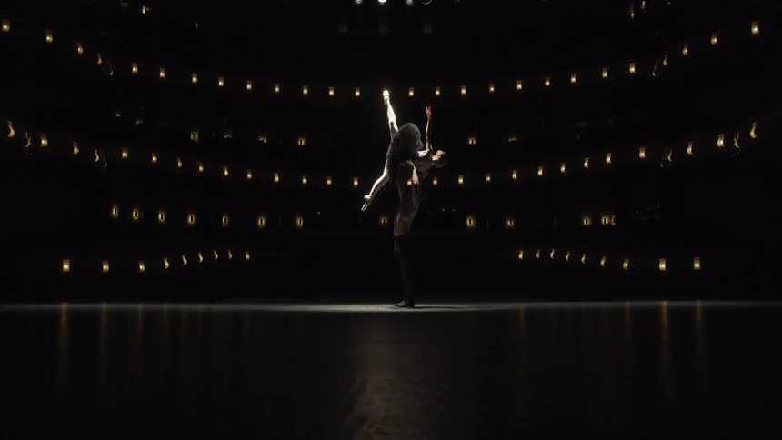 A male dancer lifts a female dancer on a dimly lit stage her leg extended gracefully