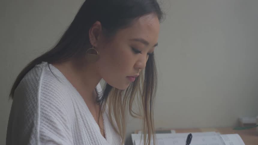 A person with long dark hair appears to be writing or drawing at a desk in a room