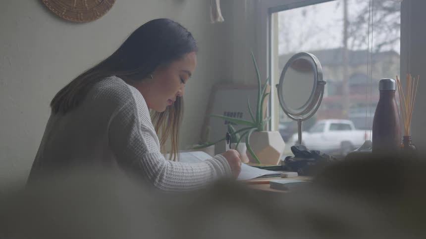 A person with long dark hair writing or drawing at a desk in a room with a window overlooking an overcast outdoor scene