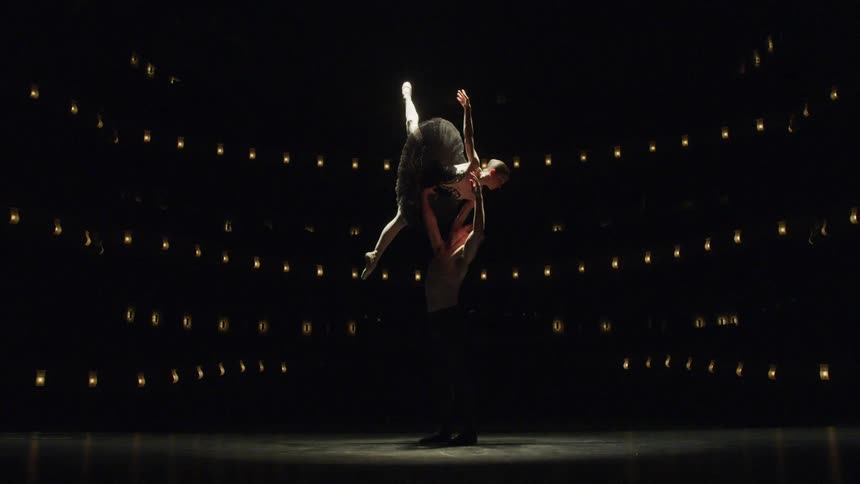 A male dancer lifts a female dancer on a dimly lit stage her leg extended gracefully