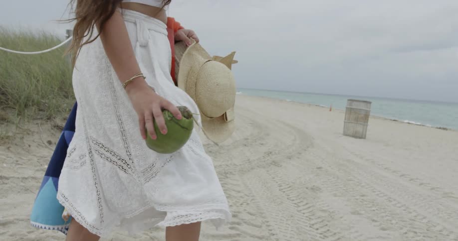 A woman carrying a hat and towel walks on the sand towards the ocean