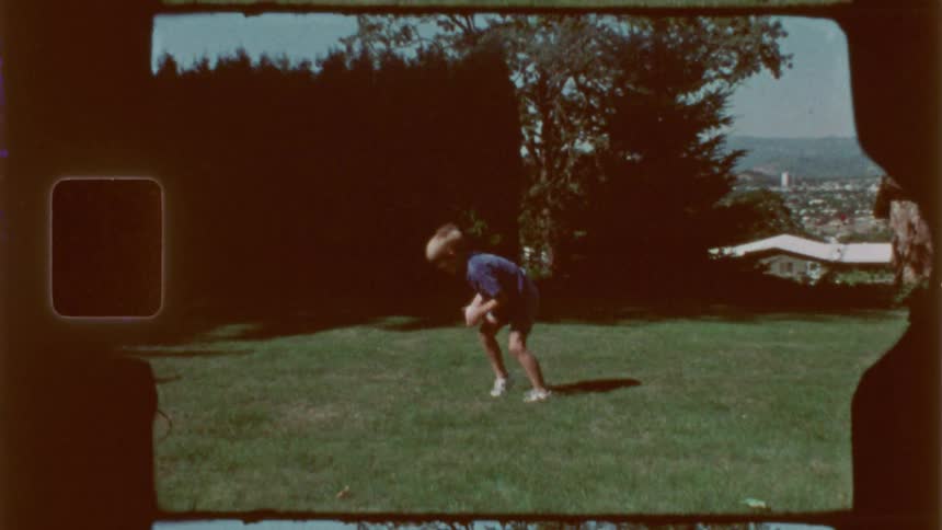 A vintagestyle shot shows a child in shorts and a tshirt playing with a ball on a grassy lawn with trees and a building in the background
