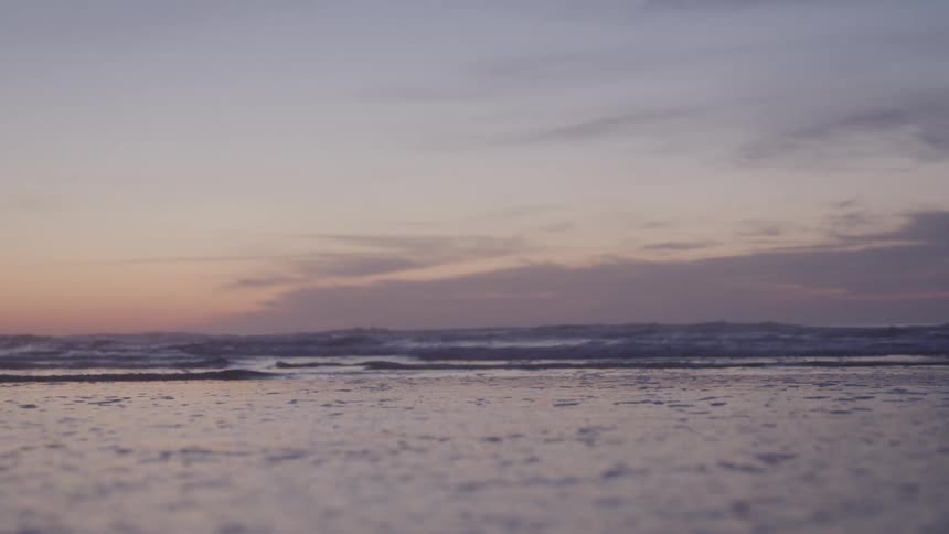 From a low vantage point the intricate texture of the oceans surface is prominent as gentle waves rhythmically roll towards the sandy shore meeting the distant horizon under a soft diffused sky