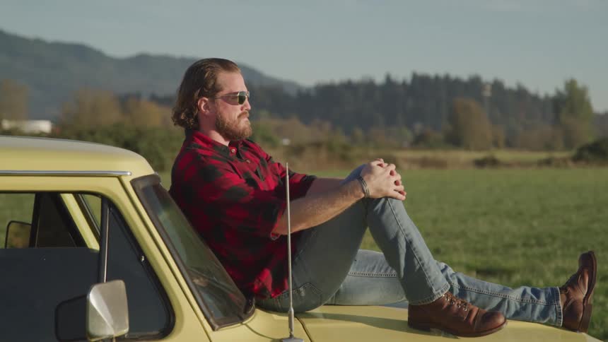 Man wearing sunglasses sitting on the hood of a car leaning his back against the windshield and enjoying the view of a green field
