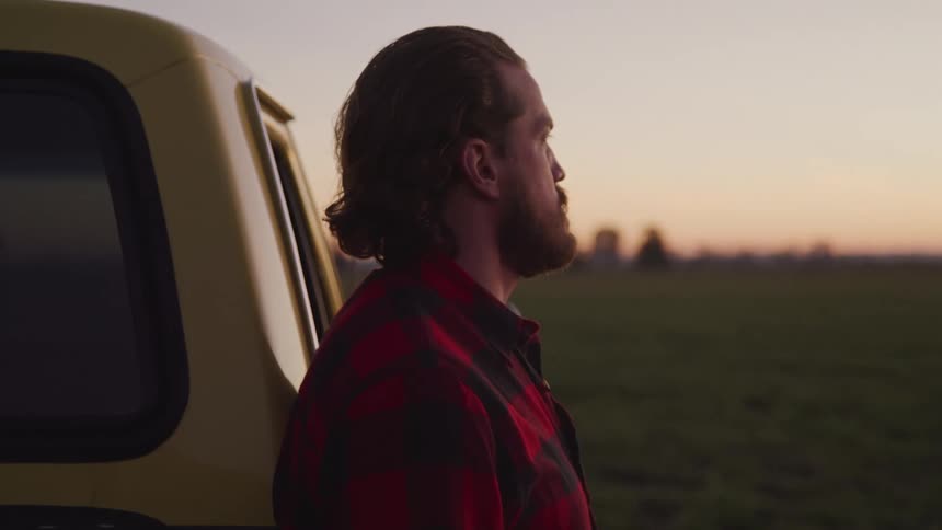 A person with a beard and long hair wearing a red plaid shirt looks off into the distance at a sunset landscape reclined on a yellow vehicle