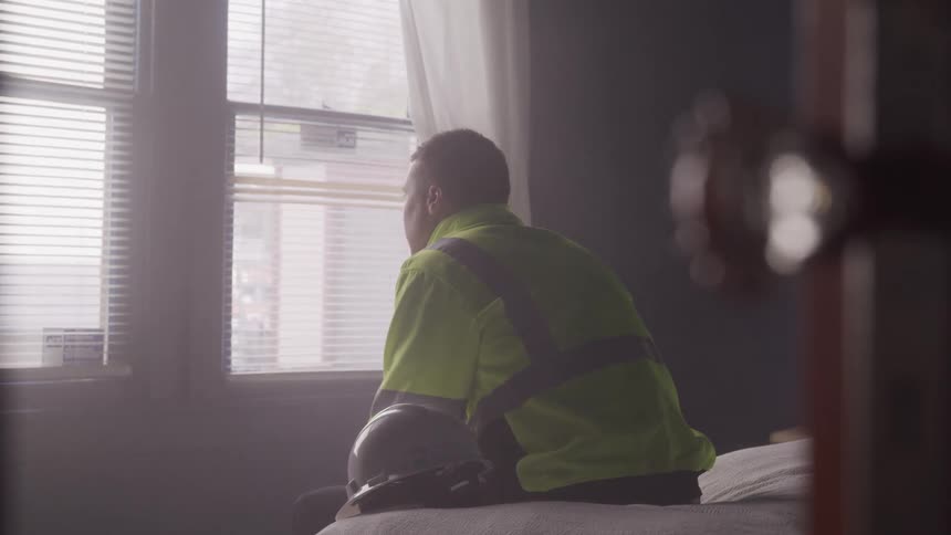 A person in a highvisibility jacket sits on a bed gazing out a window with partially open blinds while a hard hat rests beside him
