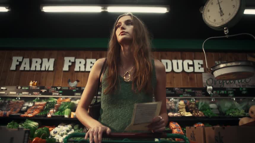 A young woman with long hair is pushing the shopping cart through the produce center as she is reading her grocery list