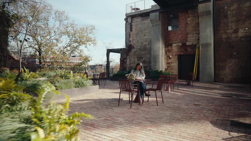 A person sits at a table in an outdoor courtyard surrounded by weathered brick structures and foliage