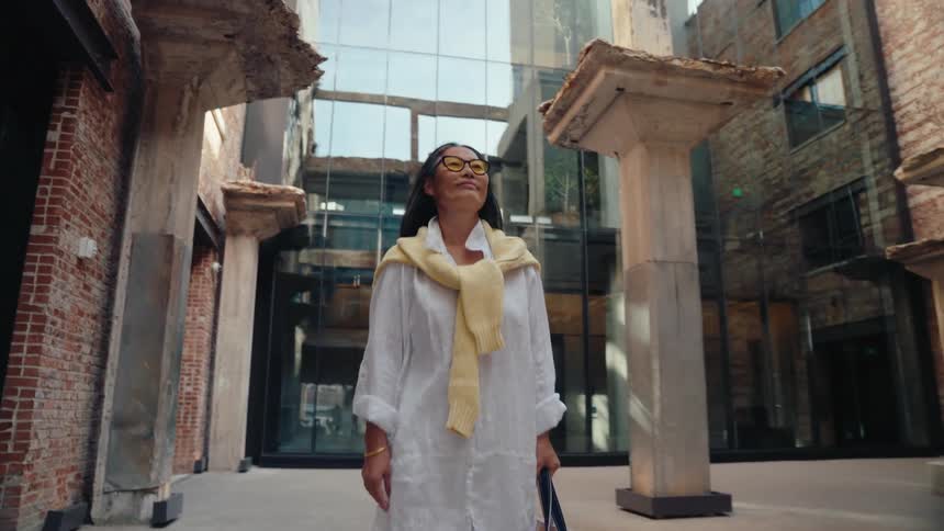 A woman in a lightcolored outfit walks around in a courtyard surrounded by weathered columns and modern buildings looking upwards