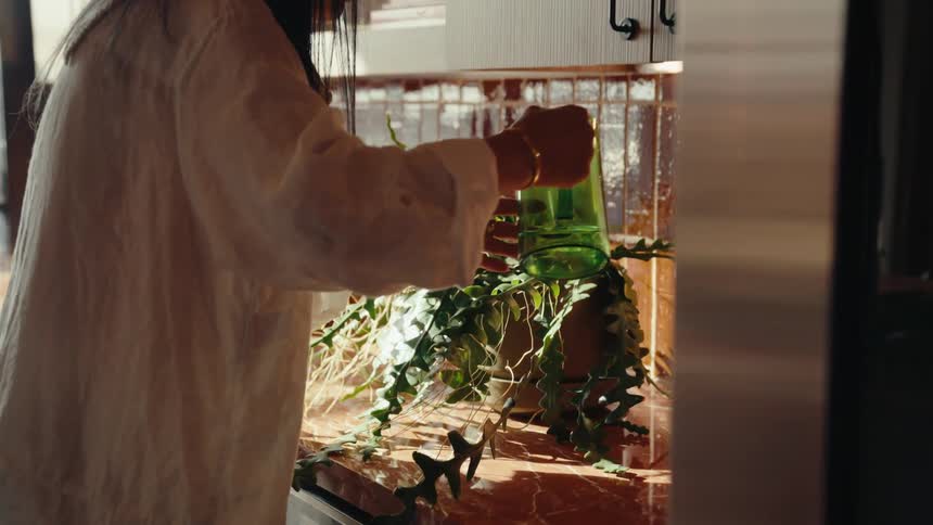 Woman filling a jug with water in the kitchen sink and then pouring it into a potted plant
