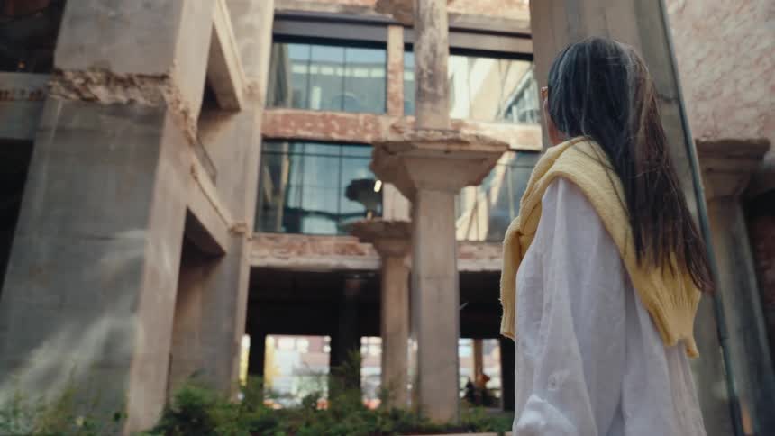 A woman in a lightcolored outfit walks around in a courtyard surrounded by weathered columns and modern buildings looking upwards