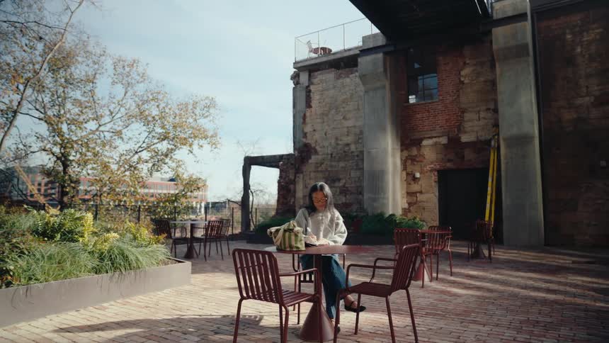Woman sits at an outdoor table near a large building and writes in a notebook