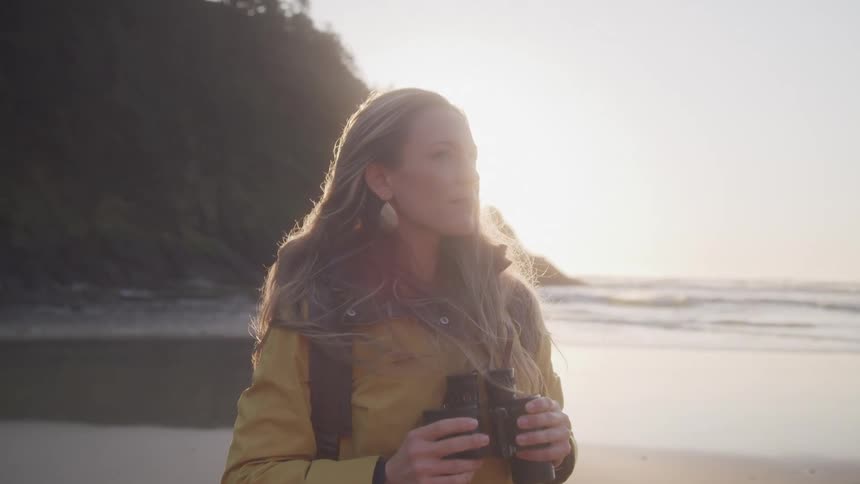 Woman stands on the beach with a pair of binoculars back lit by the sun shining past a nearby forested hill