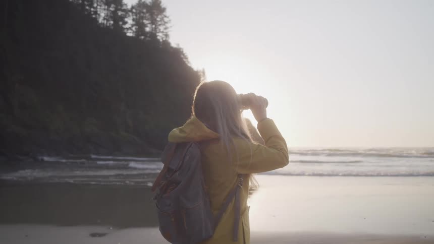 Woman with a backpack and using binoculars on a beach at dawn
