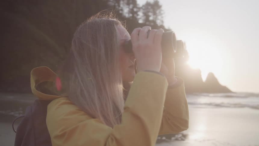 Woman using binoculars while standing on a beach with the sun on the horizon