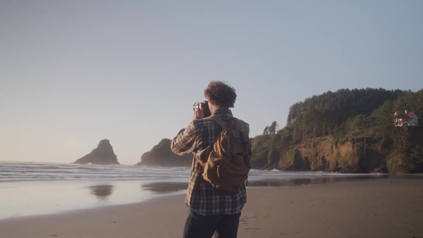 A man is using a camera as he stands on a rocky beach surrounded by cliffs