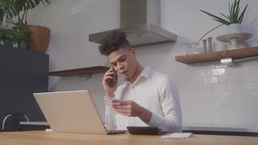 A man is talking on a cellphone as he stands in front of a table with a laptop
