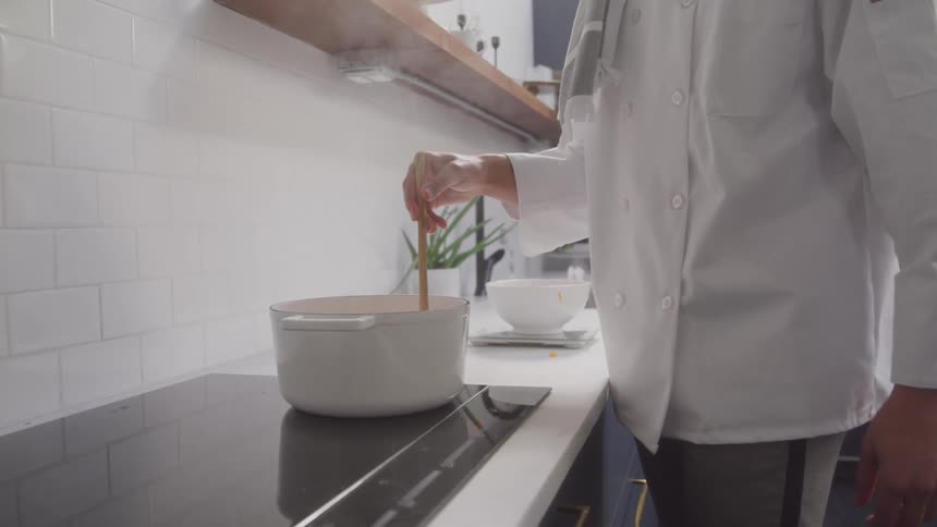 A chef wearing a white coat is actively stirring the contents of a white pot placed on a black electric stovetop using a wooden ladle as a utensil in a kitchen environment with white tiled walls