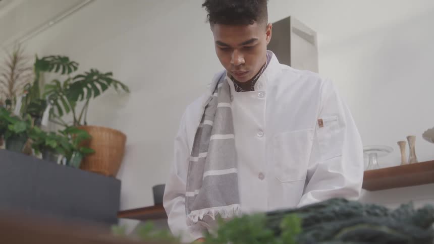 From a low angle a chef in a white uniform is intently focused at a table preparing food thats just below the cameras view with plants visible in the background