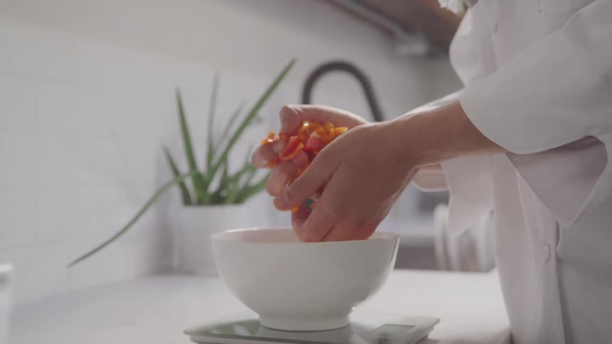 A woman dressed in white is handling some chopped fruit from a bowl placed on a scale on the kitchen counter