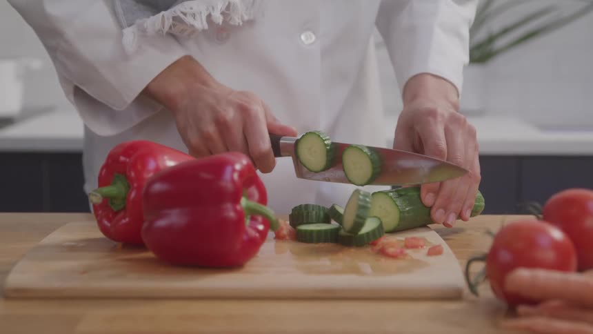 Hands are shown precisely slicing cucumbers into rounds with a sharp knife on a wooden cutting board with red bell peppers and other vegetables placed around the cutting surface ready to be prepared