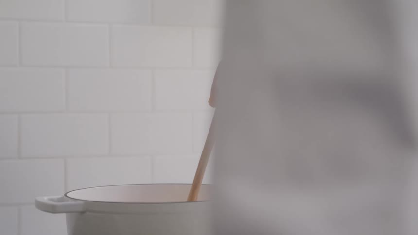 A persons hand stirs something in a white pot with a wooden spoon against a backdrop of white tiled walls