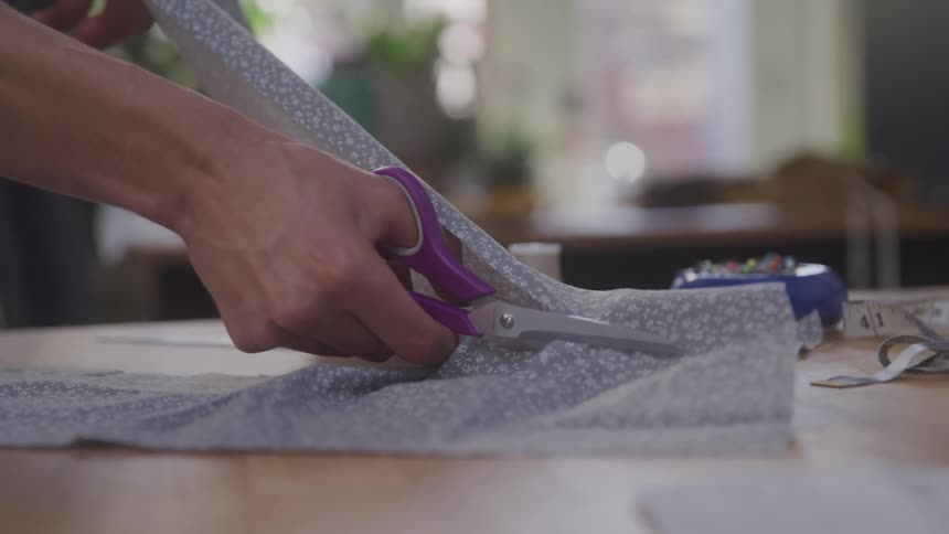 With focused precision hands guide a pair of scissors as they smoothly cut through patterned fabric with measuring tape and a needle cushion resting nearby on the surface