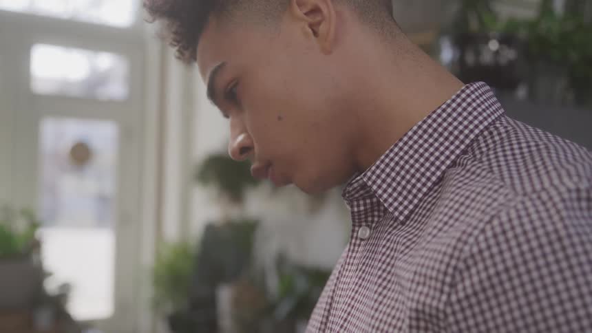 A young man wearing a checkered shirt stands indoors with plants and a window in the background