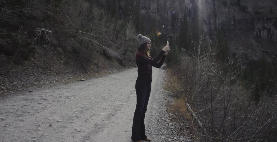 A woman stands on the side of a dirt road taking a picture of the scenery