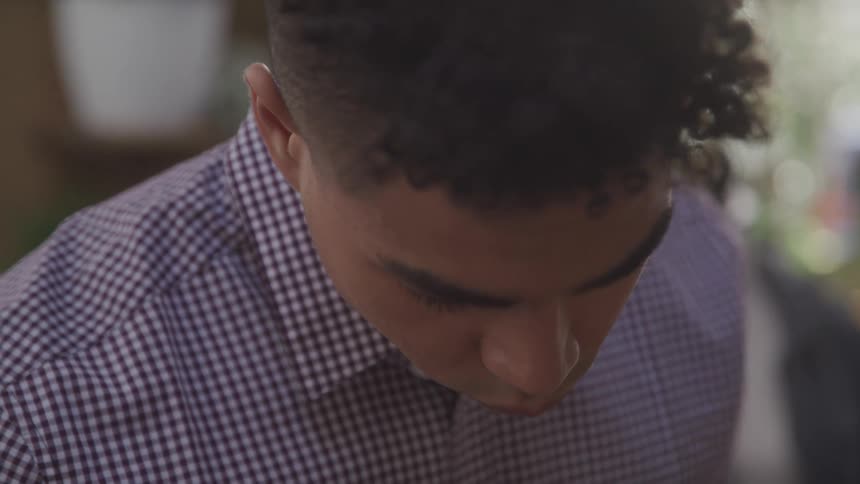 A man with curly hair glances downwards his face softly lit wearing a checkered shirt in a closeup shot highlighting his lowered gaze and relaxed demeanor