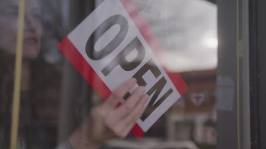 A woman approaches the glass door of a shop to change the sign actively switching it from closed to open signaling a change in business status as seen from outside the entrance