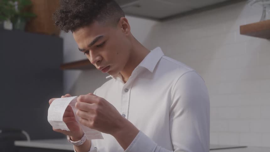 Young man looking concentrated at a receipt he holds in his hands