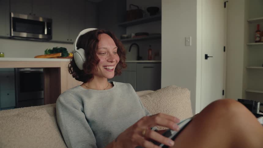 A woman with headphones sits on a couch looking at a book or magazine in her hands