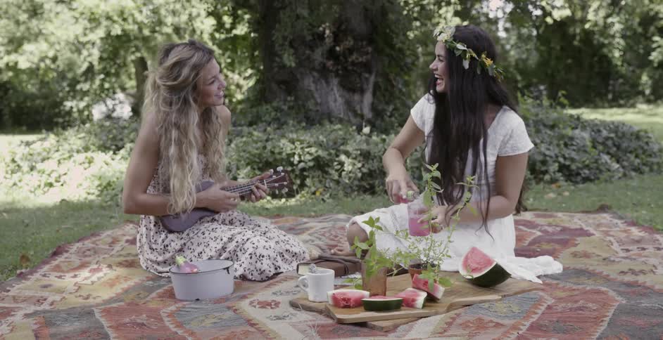 Two women enjoying an outdoor picnic as one pours a drink while the other is playing a ukulele