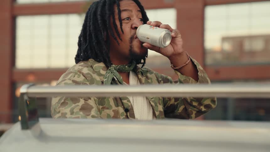 A man with dreadlocks wearing a camouflage jacket grills food on a balcony while drinking beer with an urban cityscape in the background