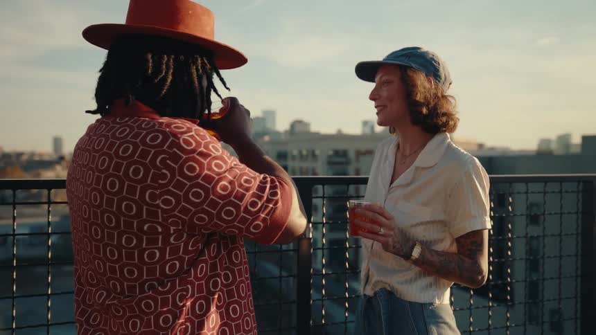 A woman with tattoos and a cap holds a drink while talking to a man in a hat on a rooftop with a cityscape in the background