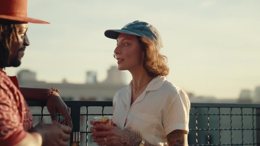 A woman with tattoos and a cap holds a drink while talking to a man in a hat on a rooftop with a cityscape in the background