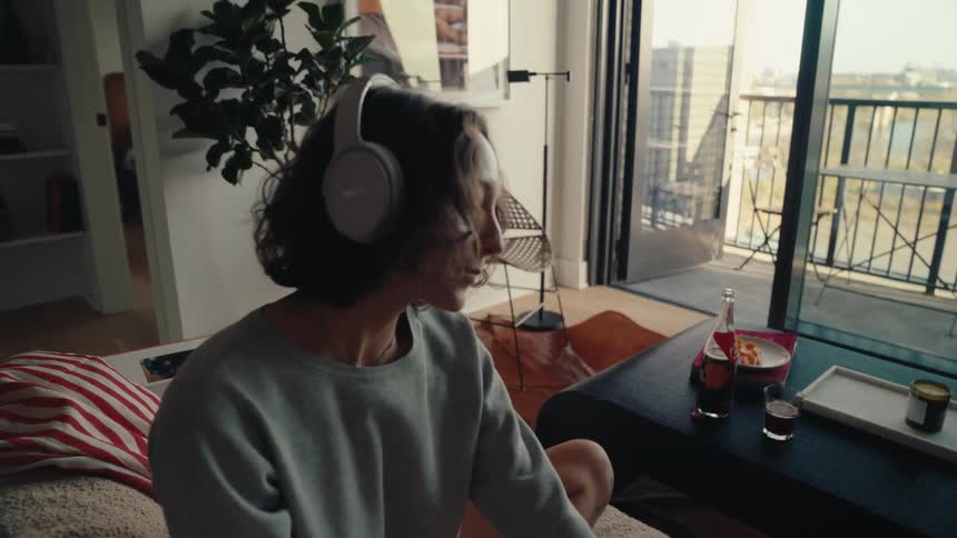 Woman sitting on the sofa in the living room putting on headphones and eating food