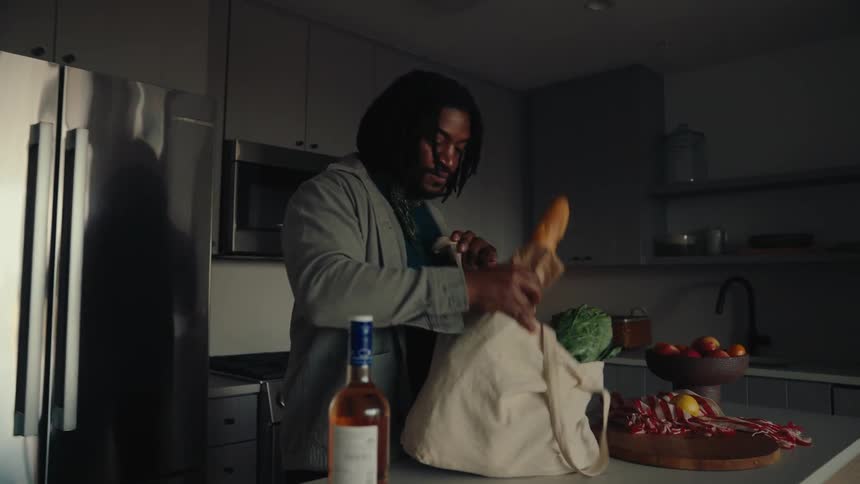 Woman sitting on the couch with headphones and in the background a man arrives with a shopping bag with vegetables and a wine