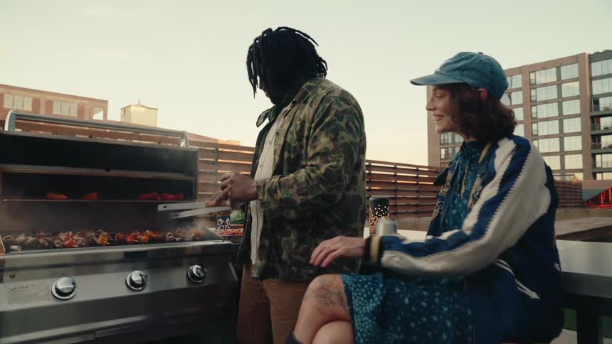 Man roasting meat while being accompanied by a woman who is sitting next to him