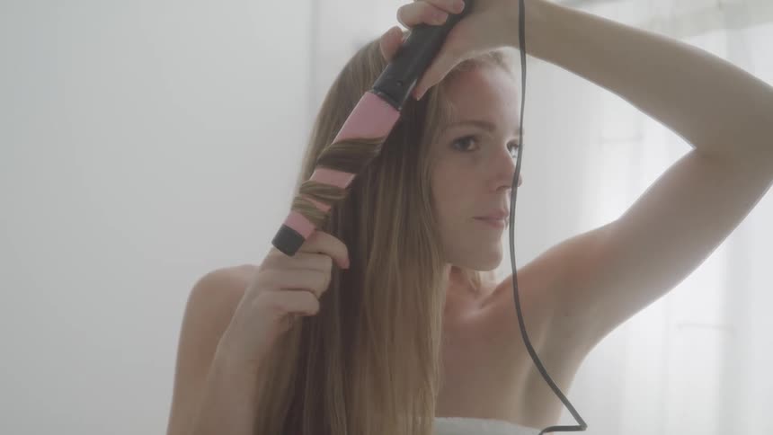 A young woman is curling her long hair using a curling iron in front of a bathroom mirror