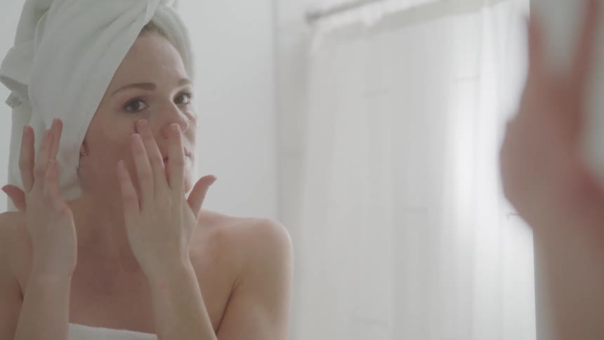 A woman wearing a towel on her head applies moisturizer to her face while looking in the bathroom mirror