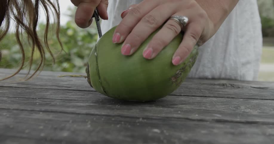 A woman is using a knife to cut a coconut on a picnic table